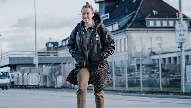 Eine VfL-Wolfsburg-Spielerin geht über die Straße. Sie trägt einen schwarzen VfL Mantel, ihre Hände sind rechts und links jeweils in der Jackentasche. 
