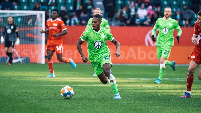 Ridle Baku im VfL Wolfsburg Sondertrikot mit Peace Zeichen läuft mit dem Ball am Fuß über das Spielfeld, im Hintergrund gegnerische Spieler von Union Berlin.