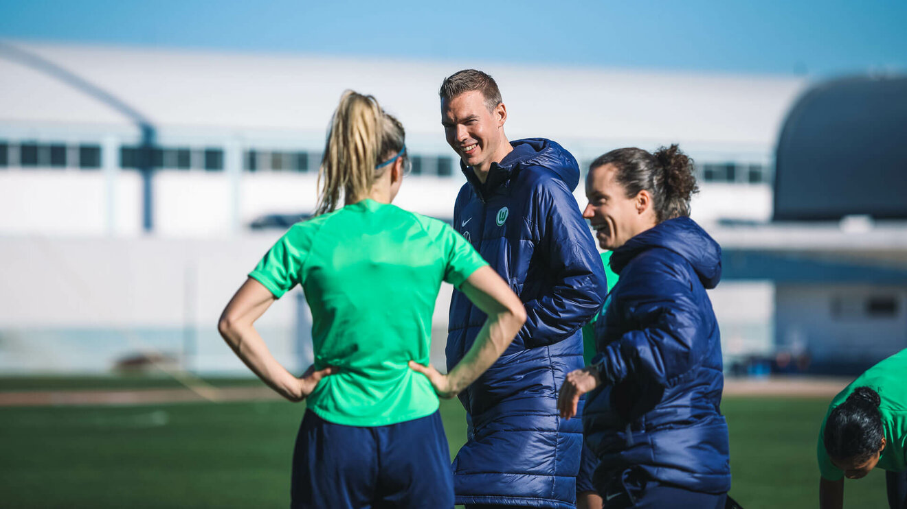 Der VfL Wolfsburg-Trainer Tommy Stroot steht lachend auf dem Trainingsplatz neben Sabrina Eckhoff.