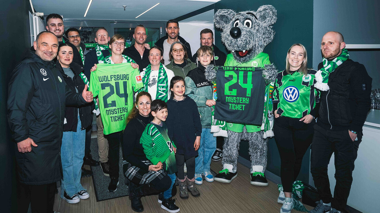 VfL-Wolfsburg-Maskottchen Wölfi steht zwischen Fans die Trikots hochhalten.