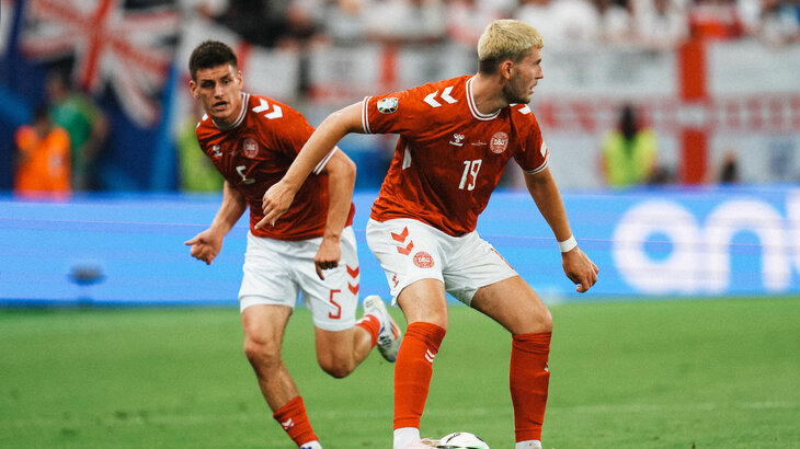 Die VfL-Wolfsburg-Spieler Joakim Maehle und Jonas Wind im Trikot der dänischen Nationalmannschaft. Beide sind in einem Stadion, während Jonas Wind den Ball am Fuß hat und Joakim Maehle hinter ihm läuft.