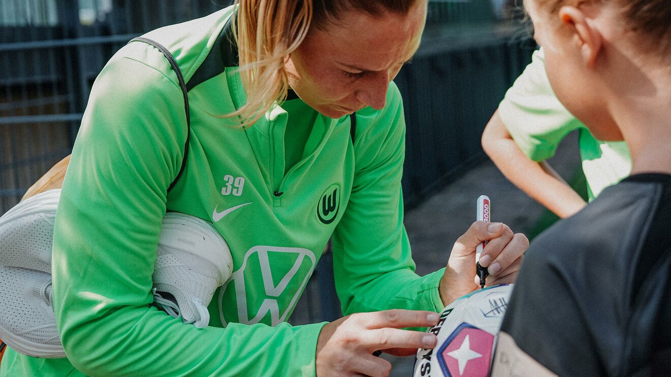 Die VfL-Wolfsburg-Spielerin Sarai Linder mit unter den Arm geklemmten Schuhen und einem Stift in der Hand. 