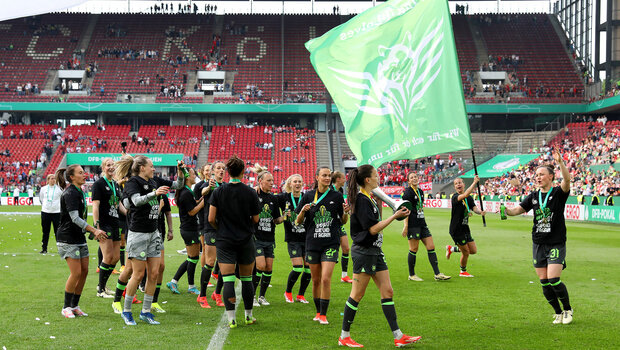 DFB-Pokalfinalle. Die Wölfinnen lassen sich auf dem Platz feiern. Sie jubeln vor Freude. Eine Spielerinn schwenkt eine Fahne in die Luft. 