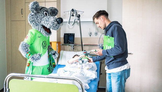 VfL-Wolfsburg-Spieler Kilian Fischer besucht gemeinsam mit Wölfi das Kinderkrankenhaus. Er übergibt einem Patienten ein Wölfi-Kuscheltier.