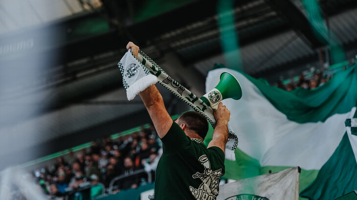 Ein VfL-Wolfsburg-Fan hält einen Schal und ein Megafon in den Händen.