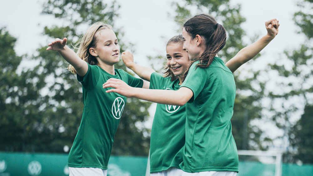 Drei Mädchen in VfL-Wolfsburg-T-Shirts stehen auf einem VfL-Trainingsplatz und umarmen sich.