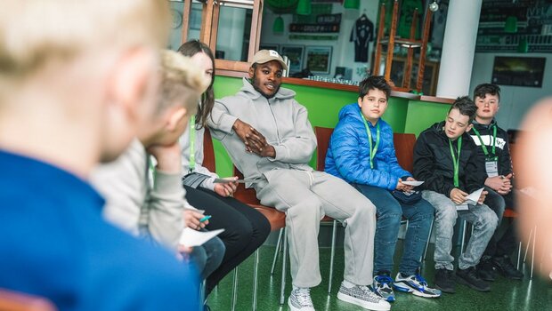 Der Profifußballer Ridle Baku sitzt mit Kindern beim Zukunftstag des VfL Wolfsburg im Stuhlkreis und erklärt ihnen seinen Beruf.