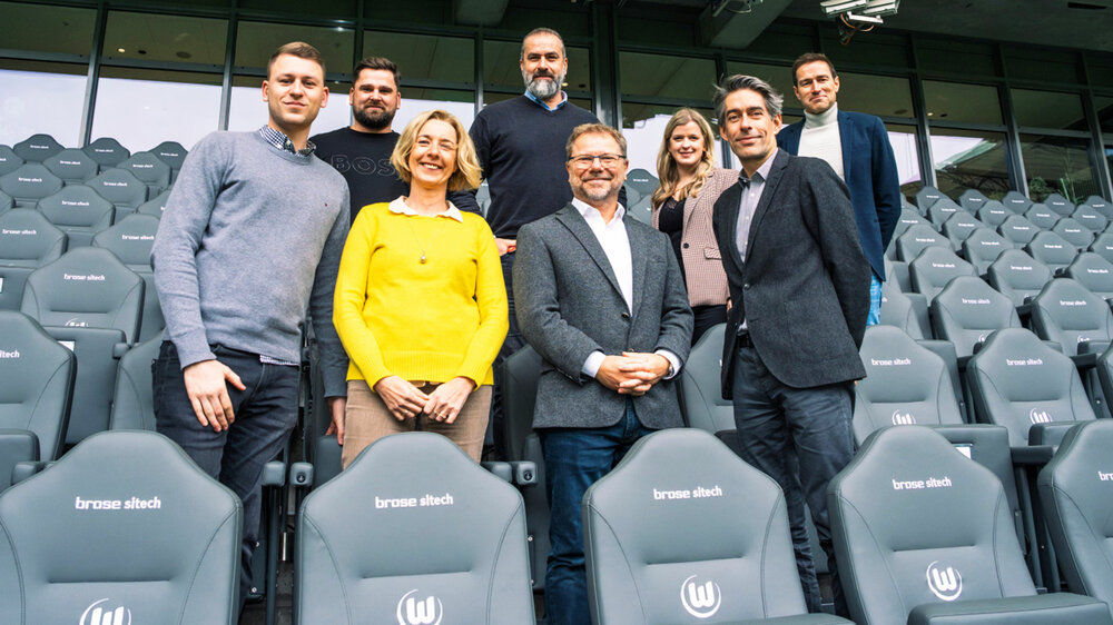 Die Verantwortlichen von Brose Sitech und seitens des VfL Wolfsburg stellen sich für ein Gruppenfoto zwischen den grauen Sitzmöglichkeiten auf.
