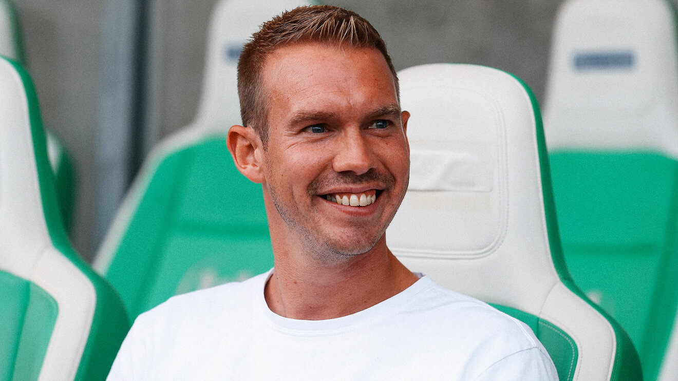 Der VfL-Wolfsburg-Trainer Tommy Stroot sitzt lächeln auf der Trainerbank.