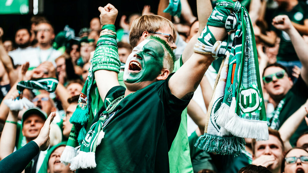 Nahaufnahme eines Fans des VfL Wolfsburg in der Nordkurve, er hat ein grün-weiß bemaltes Gesicht und mehrere Schals an den Armen, die er freudig in die Luft streckt.
