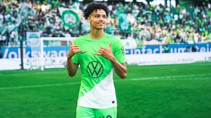 Kevin Paredes vom VfL Wolfsburg macht ein Handzeichen und grinst.
