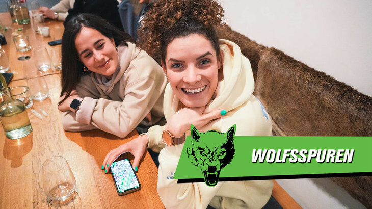 VfL Wolfsburg Spielerin Janssen sitzt am Tisch, ein Handy liegt vor ihr, und sie lacht, den Kopf auf den Hand abstützend, in die Kamera. Im Hintergrund sitzt Mitspielerin Rubio.