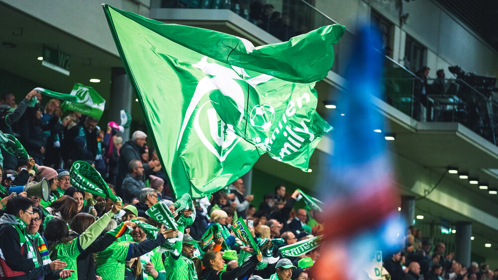 Die Fans des VfL-Wolfsburg feiern und schwenken eine große grüne Flagge.