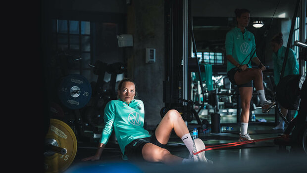 VfL Wolfsburg Torhüterin Schmitz sitzt im Fitnessstudio im Trainingslager auf dem Boden und mach mit einem Gummiband am Fuß Beinübungen.