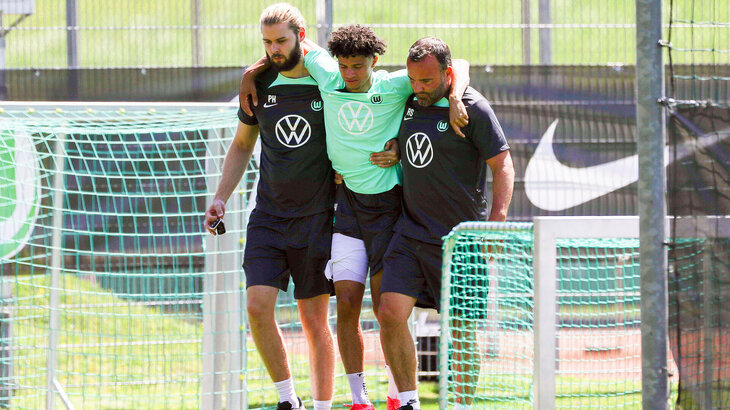 VfL Wolfsburg Spieler Kevin Paredes humpelt, gestützt von zwei Helfern, vom Spielfeld.