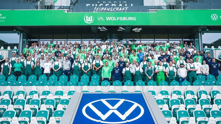 VfL-Wolfsburg-Mitarbeiter posieren in Latzhosen auf der Tribüne der Volkswagen Arena für ein Foto.