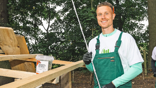 Der VfL-Wolfsburg-Spieler Yannick Gerhardt hält beim Wir für Euch-Tag einen Zollstock in den Händen und lächelt in die Kamera.