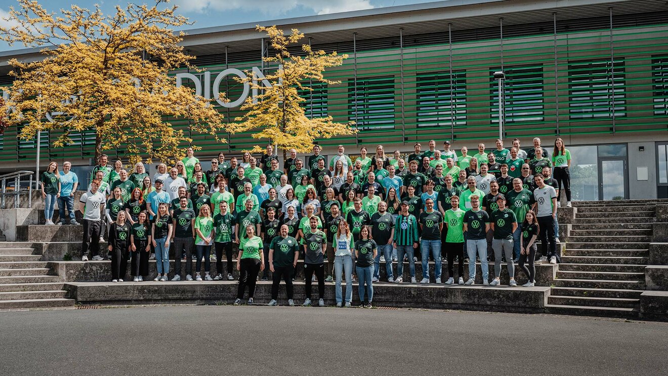 Mitarbeitende des VfL Wolfsburg tragen Trikots und stehen vor dem AOK Stadion.