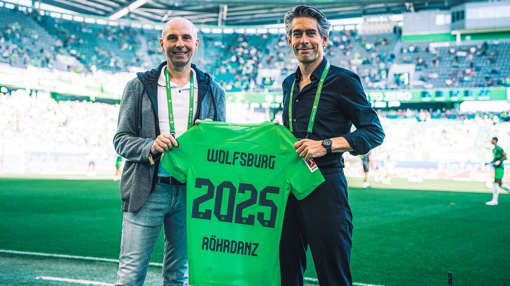 VfL-Wolfsburg-Geschäftsführer Michael Meeske und ein Partner halten gemeinsam ein Trikot mit dem Rückelflock Röhrdanz 2025 in die Kamera.