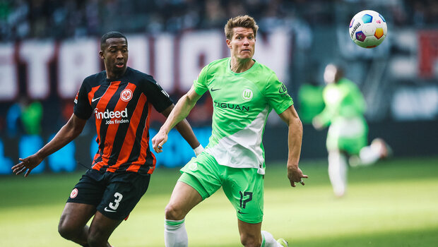 VfL-Wolfsburg-Spieler Kevin Behrens läuft mit einem Gegenspieler und fokussiert einen heranfliegenden Ball an.