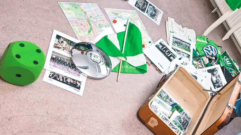 Ein Koffer und verschiedenste VfL-Wolfsburg-Artikel wie Flyer, Zeitungen, Bilder oder Trikots liegen auf dem Boden.