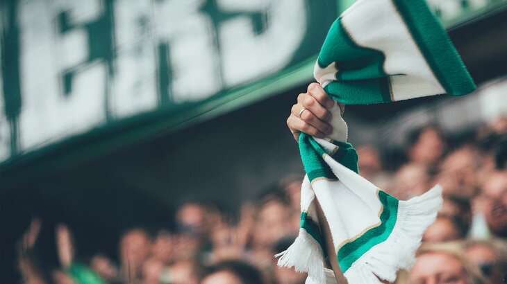 Ein Fan des VfL-Wolfsburg hält einen grün-weißen Schal in der Hand und lässt ihn kreisen.