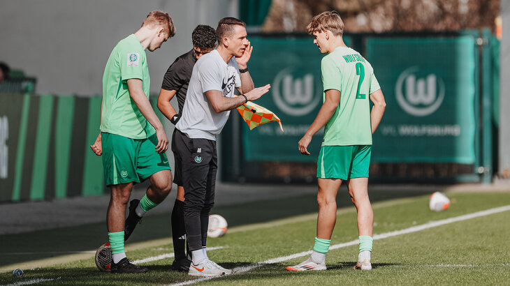 Daniel Bauer steht mit zwei Spieler der U19-Mannschaft des VfL Wolfsburg am Spielfeldrand und gibt Anweisungen.