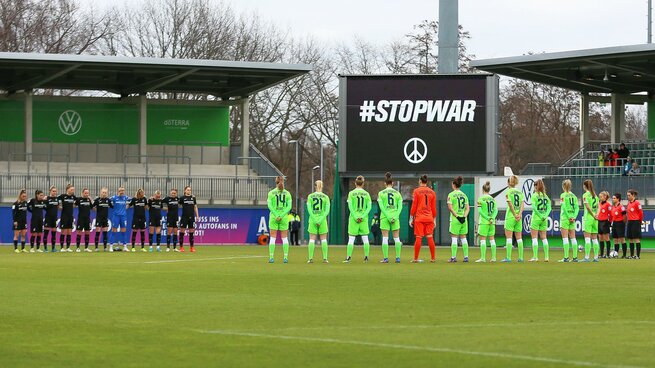 Die Frauenmannschaften des VfL Wolfsburg und SC Freiburg stehen vor Anpfiff am Mittelkreis für eine Schweigeminute - im Hintergrund zeigt die Anzeigetafel "STOP WAR" an.