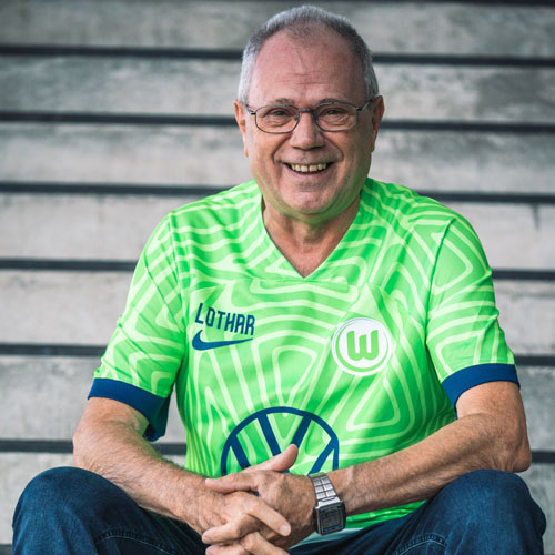 Der Fanbeauftragte Lothar sitzt im VfL Wolfsburg-Trikot auf einer Treppe.