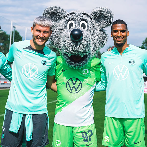 Das Maskottchen des VfL Wolfsburg, Wöfi, schaut zusammen mit Jonas Wind und Moritz Jenz in die Kamera.