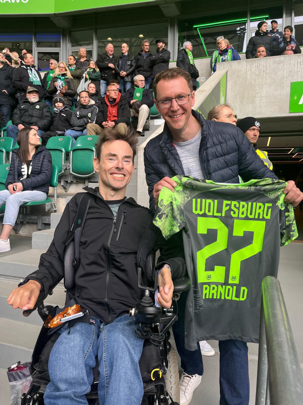 Zwei Fans des VfL Wolfsburg befinden sich am Spielfeldrand und halten ein Trikot  von Maximilian Arnold in den Händen.