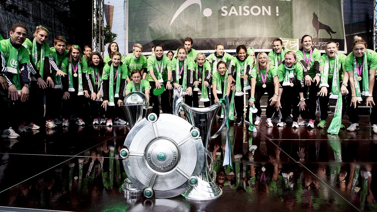 Die Frauen des VfL Wolfsburg stehen um Champions League Pokal, DFB Pokal und Meisterschale.